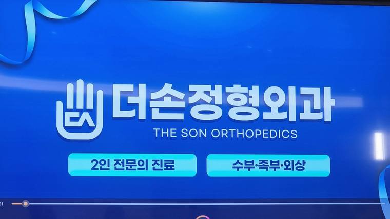 파란색 배경에 '더손정형외과'라는 문구와 영문 병원 이름, 진료 내용이 적힌 홍보용 안내판 또는 광고 화면