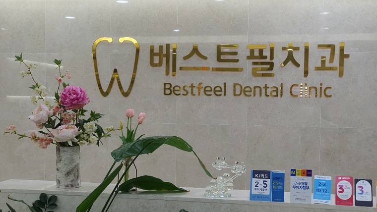 벽면에 '베스트필치과'와 'Bestfeel Dental Clinic'이라는 문구와 치아 모양의 로고가 적힌 간판이 있고, 그 앞에 꽃과 안내용 포스터들이 놓여 있다