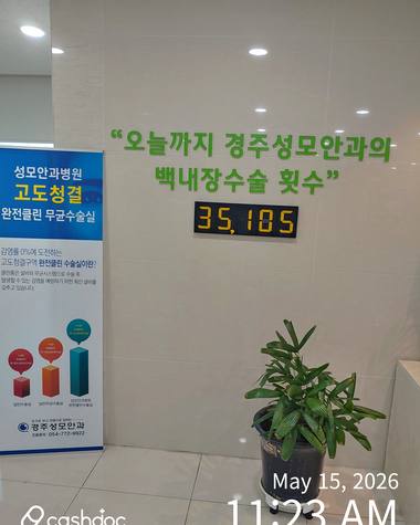 경주성모안과의원 — 벽면에 안과 홍보 문구와 가격이 적힌 포스터와 디지털 숫자가 표시된 전광판이 붙어 있으며, 그 앞에 화분이 놓여 있다