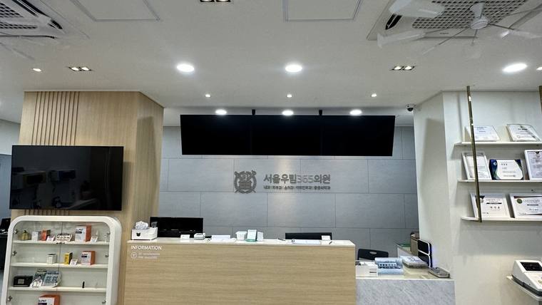 서울우림365의원 — 병원 접수대 뒤로 병원 이름이 적힌 벽면과 제품들이 진열된 선반이 보