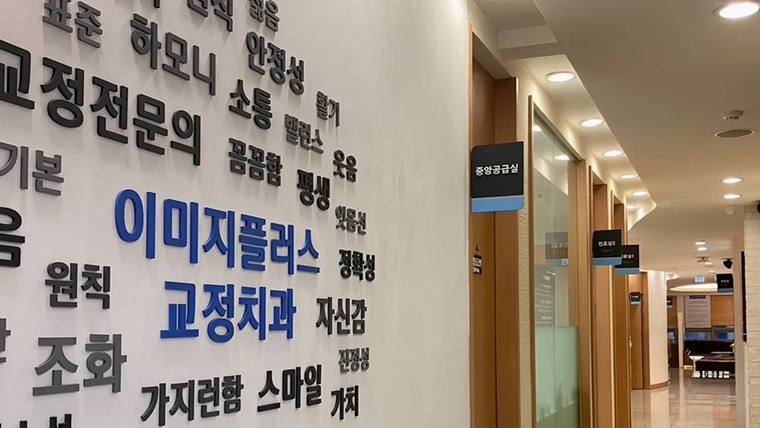 이미지플러스치과교정과치과의원 — 병원 복도 벽면에 한글 문구가 적힌 대형 안내판이 설치되어 있고 그 옆으로 진료실 입구들이 보입니다.
