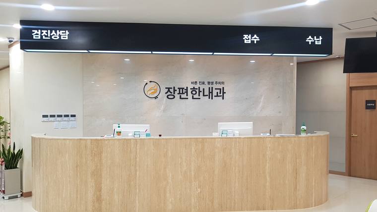 병원 접수대 위에 '장편한내과'라는 문구와 검진, 접수, 수납 안내가 적힌 간판이 있는 내부 전경
