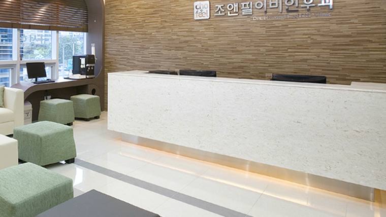 조앤필이비인후과의원 — 흰색 대리석 소재의 긴 접수대와 그 뒤로 나무 질감의 벽면이 있는 병원 내부의 로비 공간이다.